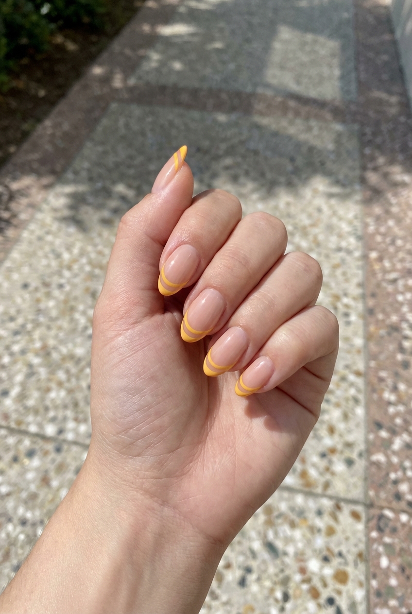 Hand slightly curled over a textured outdoor walkway displays yellow French tip nail art idea with nude glossy nails and double-layered yellow tip lines forming a striped design