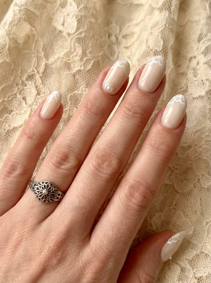 Close view of a manicured hand wearing a decorative silver ring, showing almond-shaped ivory nails with subtle lace-style white tip designs for an elegant bridal nail art idea.