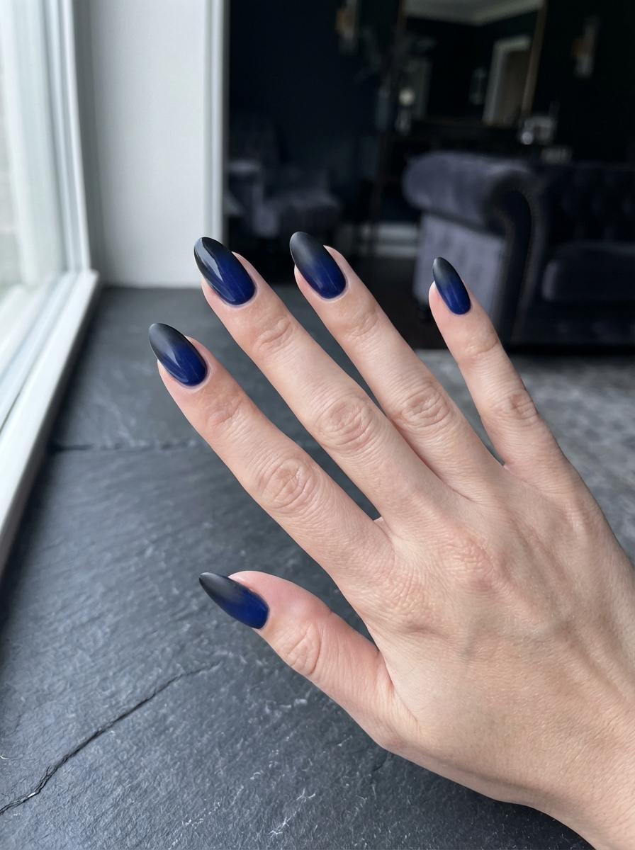 Hand placed near a window on a dark stone surface showing a black and blue nail art idea with navy blue nails fading into matte black tips on almond-shaped nails.