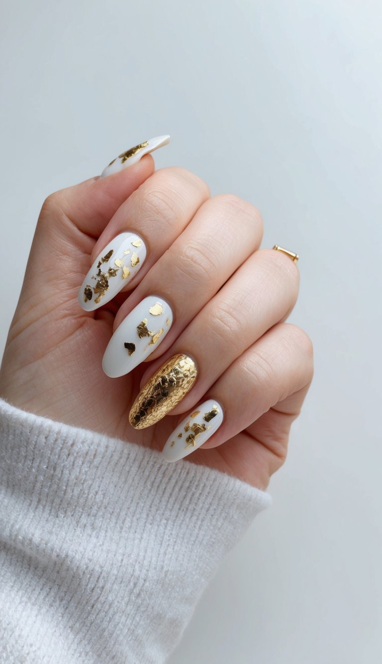 Manicured hand slightly curled against a neutral background featuring a white and gold nail art idea with white almond nails scattered with gold foil and one nail coated in textured metallic gold.
