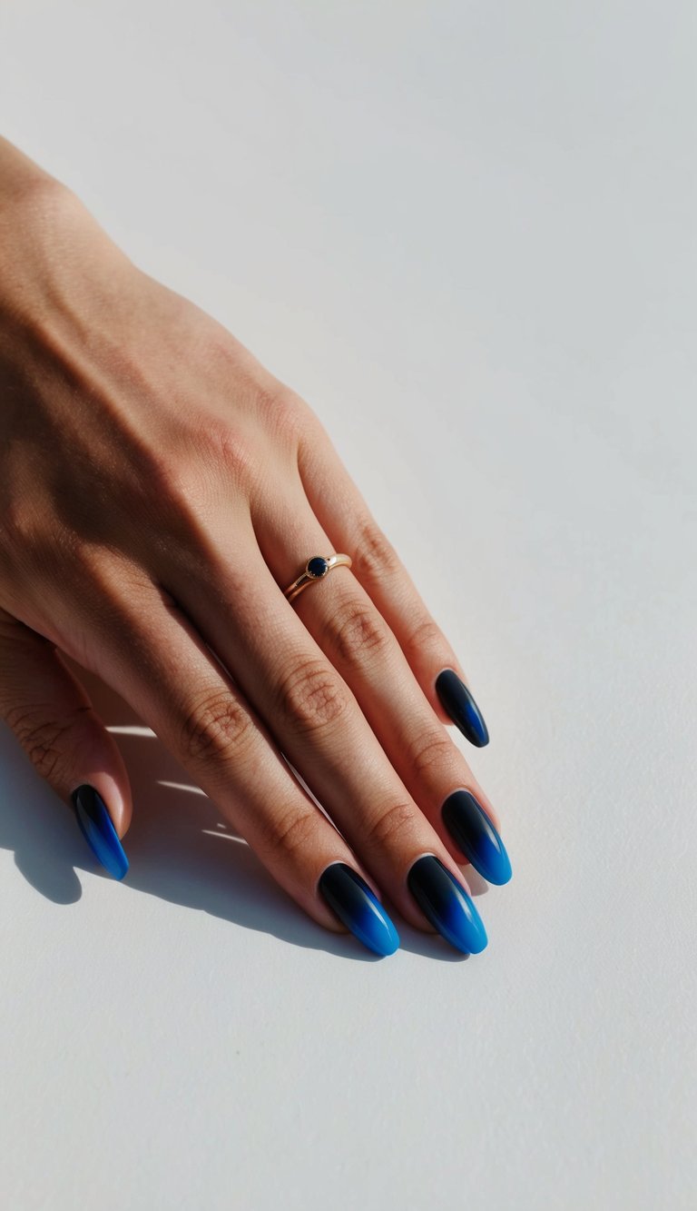 Hand resting on a bright white surface with sunlight casting shadows, showing a black and blue nail art idea featuring glossy ombré nails fading from black near the cuticle to cobalt blue tips.