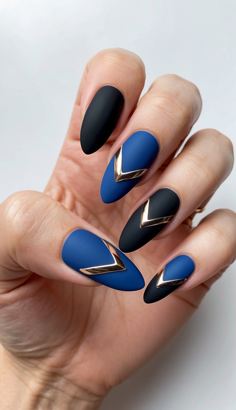 Manicured hand with almond nails displaying a black and blue nail art idea featuring matte cobalt blue and matte black polish finished with gold V-shaped metal accents.