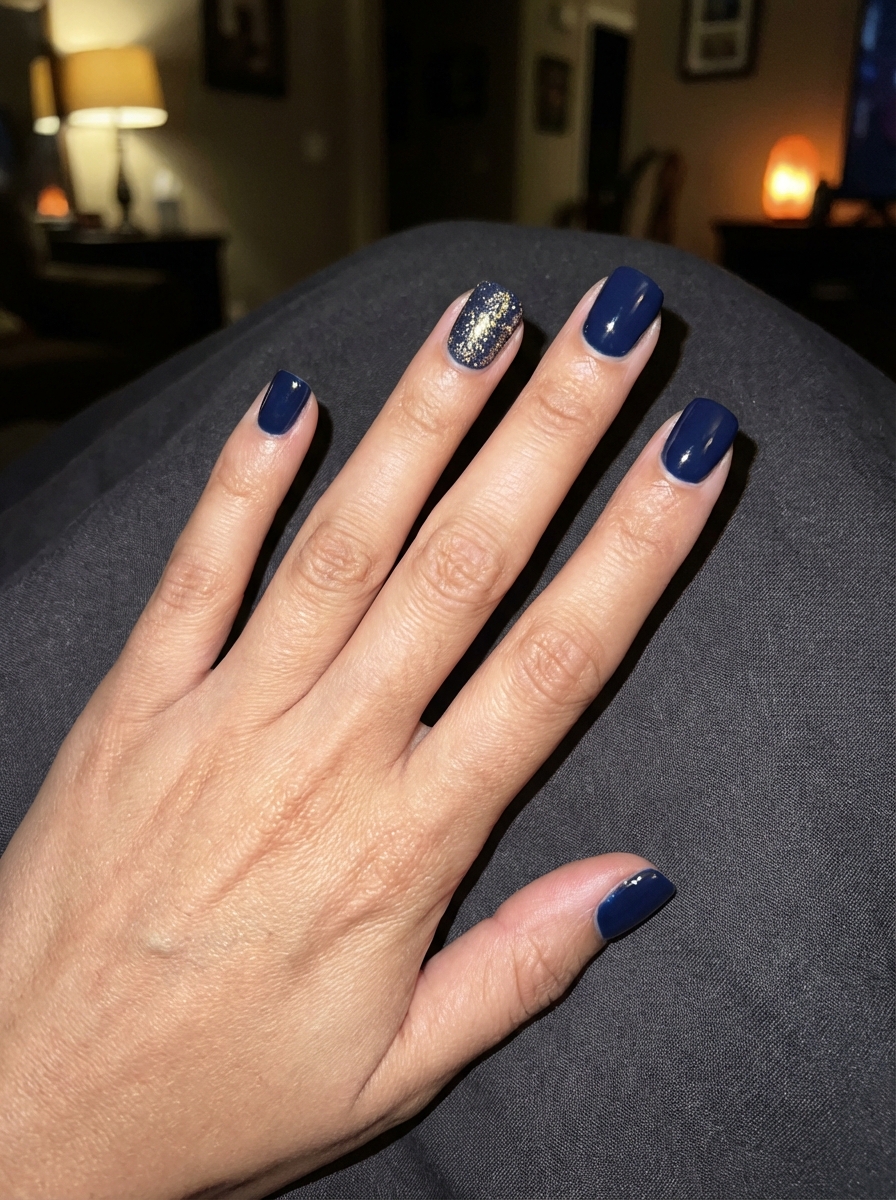 Close-up of a hand placed on dark clothing displaying a short nail art idea with dark navy blue polish and one nail covered in gold glitter on short nails.