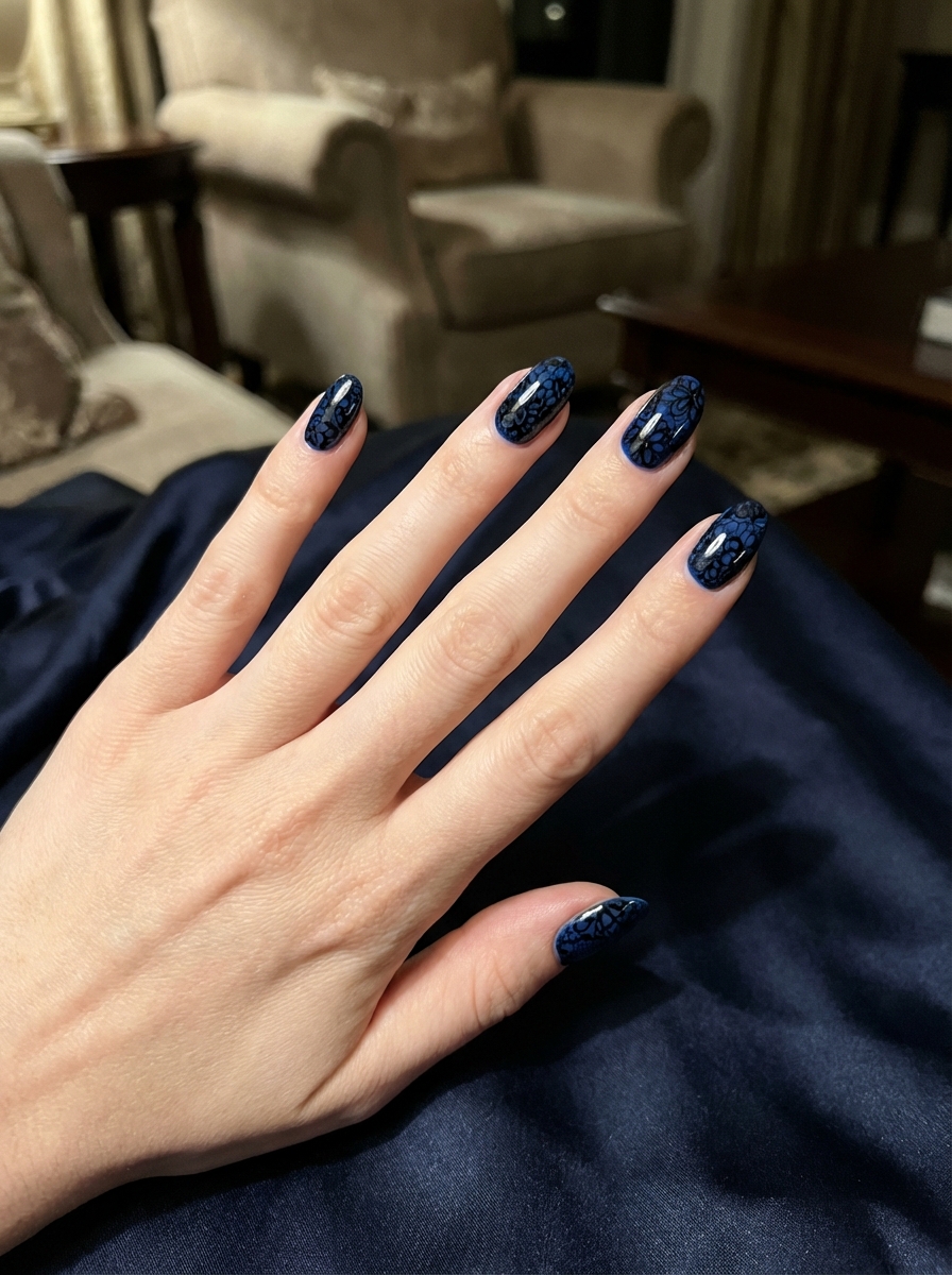 Close-up of a hand resting on dark blue fabric featuring a black and blue nail art idea with navy polish and ornate black lace-style nail art patterns.