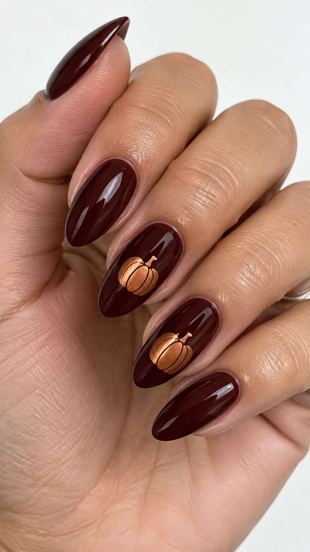 Glossy burgundy nails and small metallic copper pumpkin designs on two accent nails