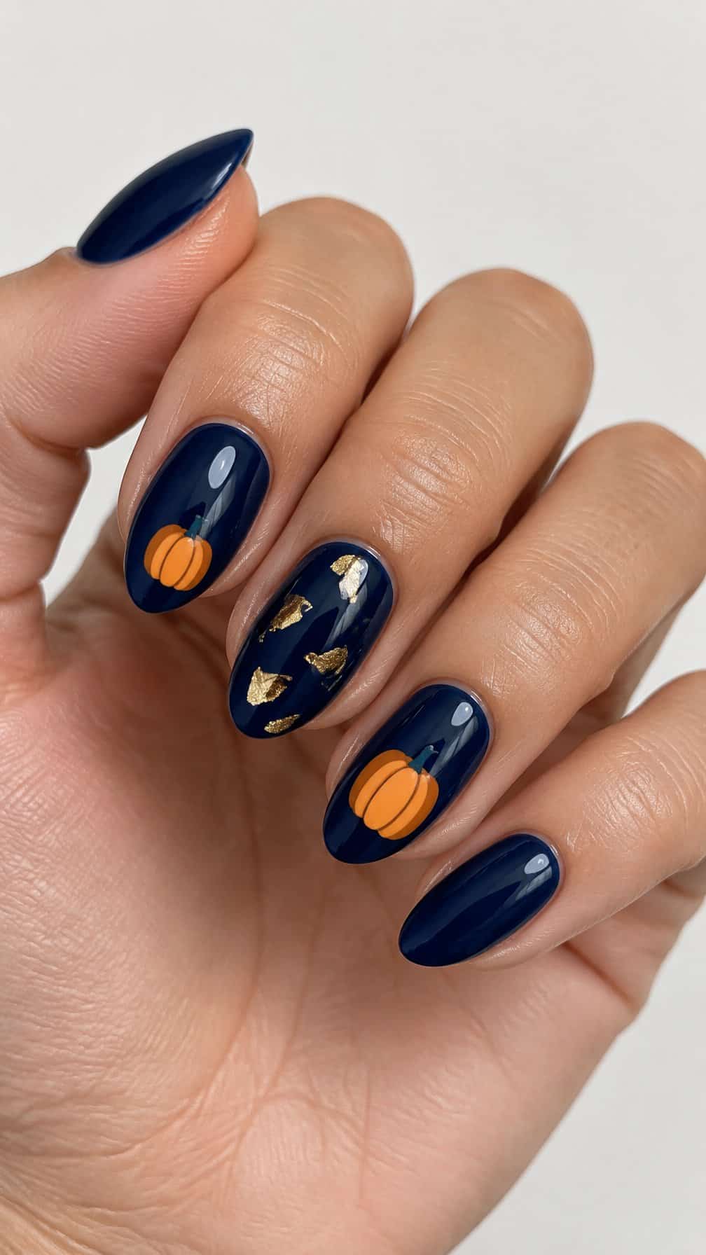 Glossy navy blue nails featuring small orange pumpkin designs and gold foil accents