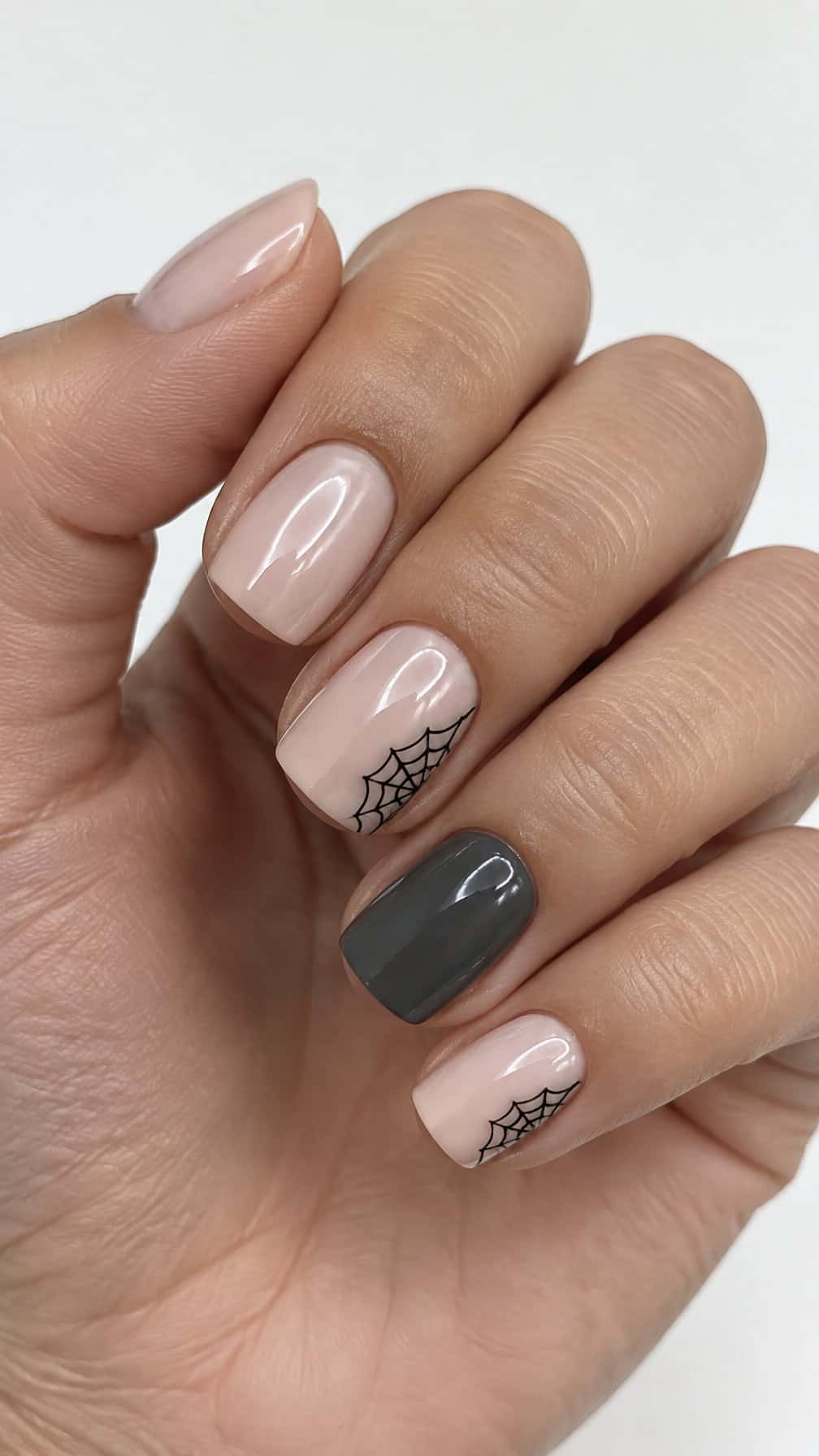 Glossy nude nails, including small black spider web designs painted in the corners of two nails