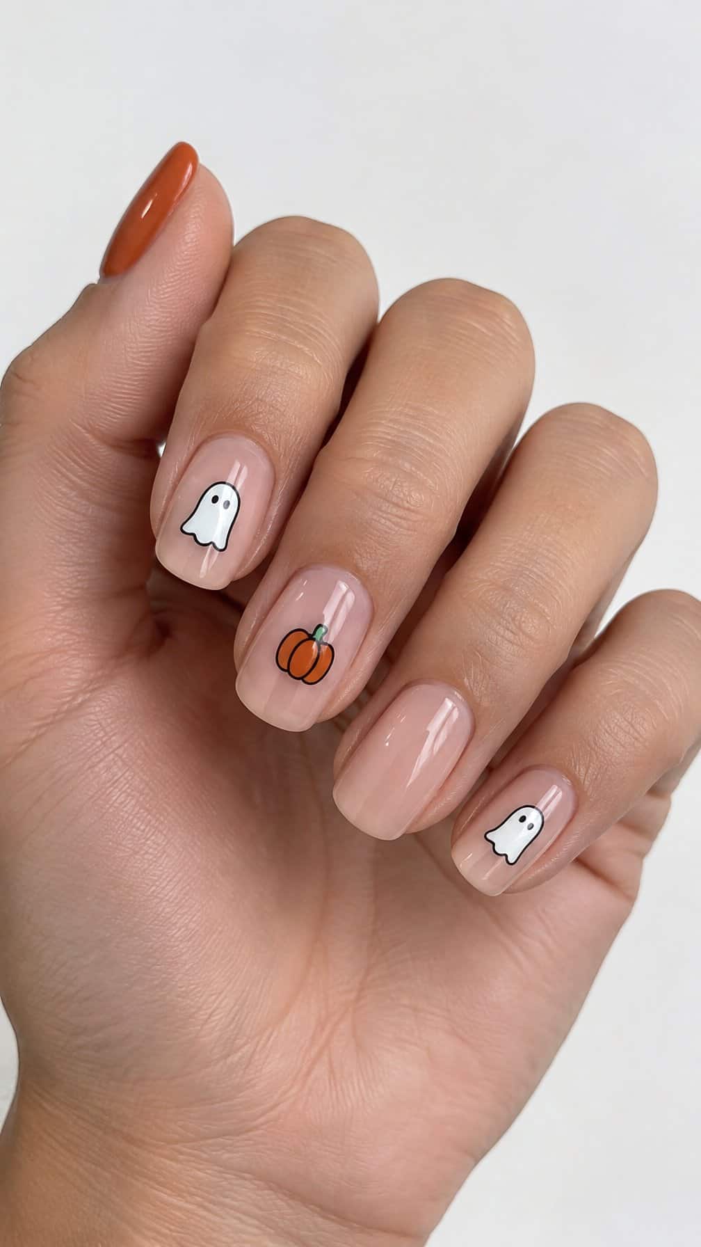 Glossy nude nails decorated with tiny white ghosts and orange pumpkins