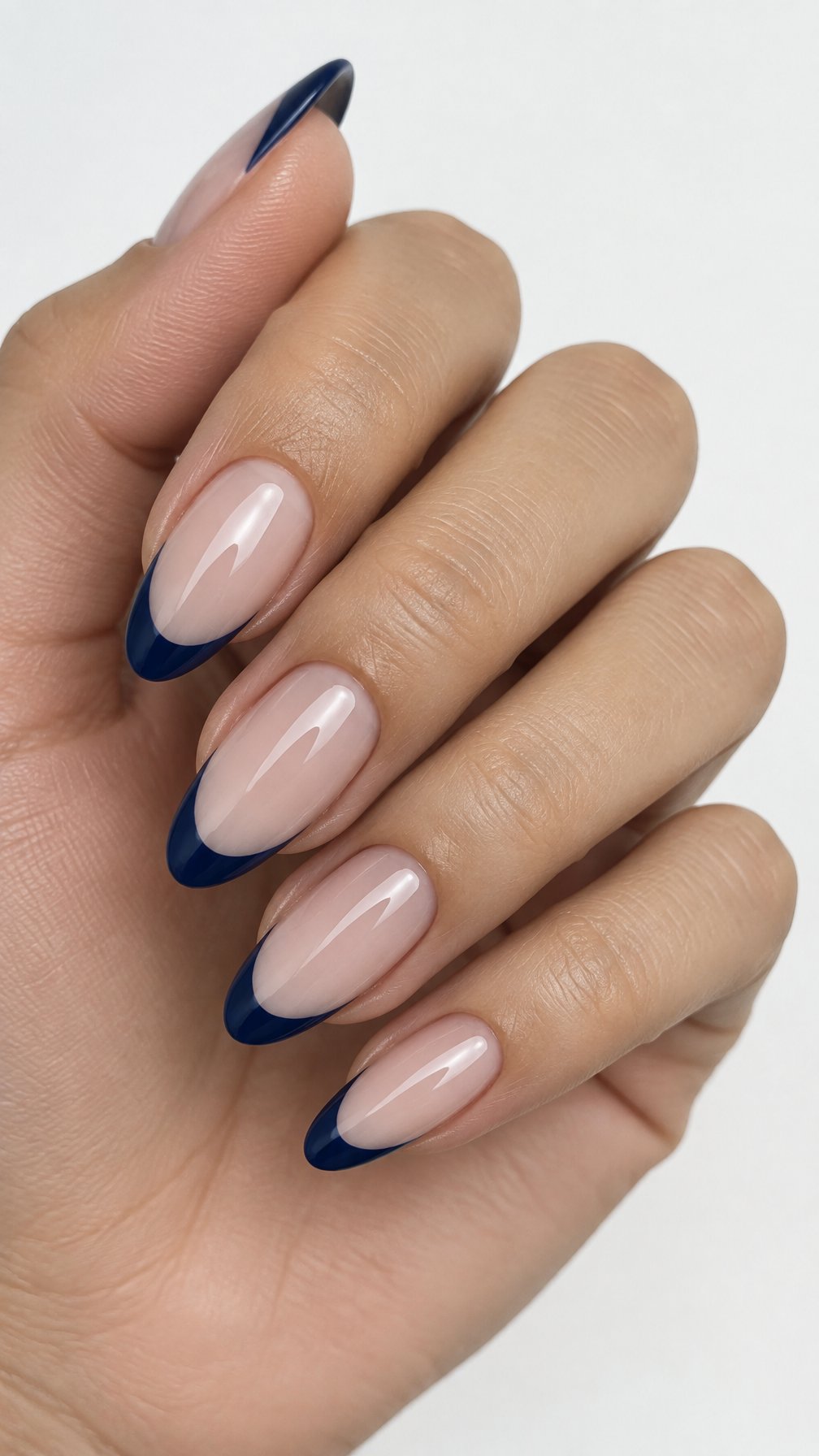 One hand with almond-shaped nails painted with sheer nude polish and midnight blue French tips against a white background.